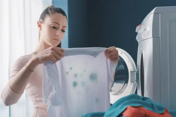 Zmartwiona kobieta trzyma białą koszulkę z zielonymi plamami, widoczne odbarwione ubrania po praniu.